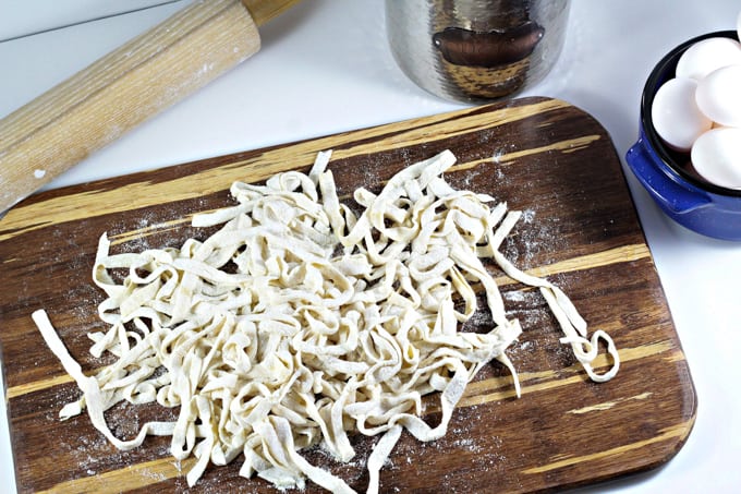Noodles on a cutting board, a bowl of eggs and a rolling pin.