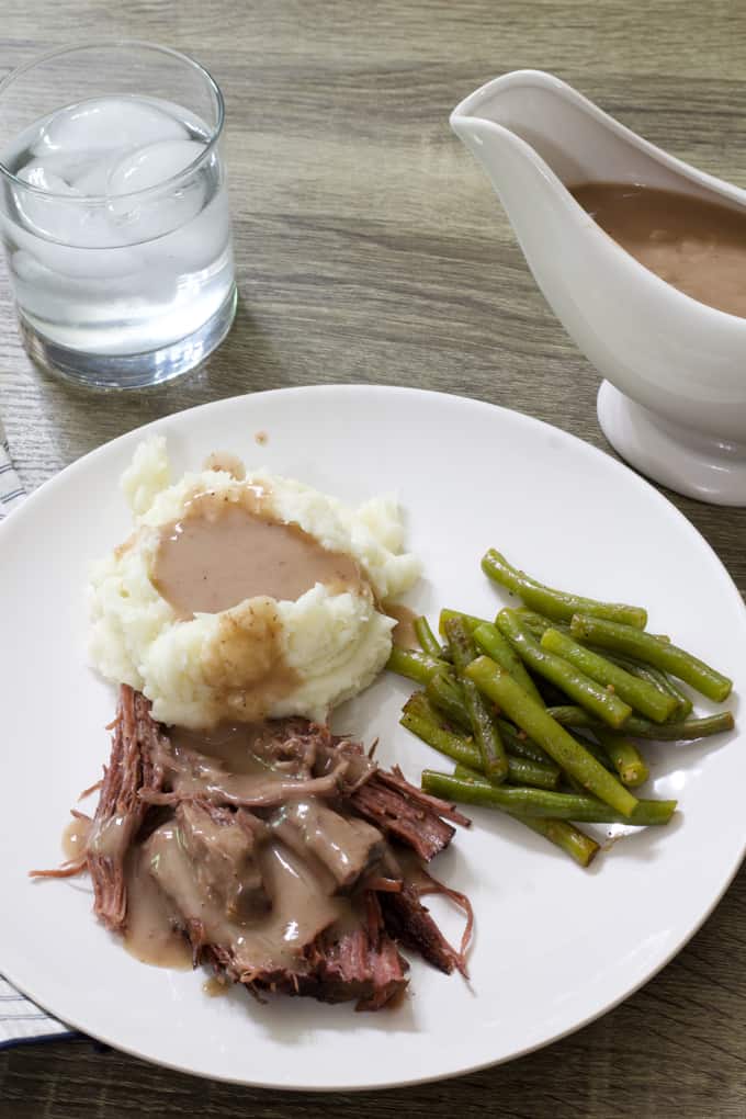 A plate of food, glass of ice water and gravy boat full of brown gravy.