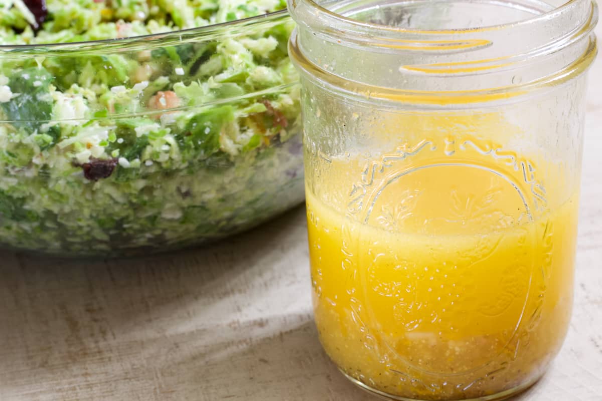 Close up view of a half full jar of citrus vinaigrette in front of a bowl of salad.