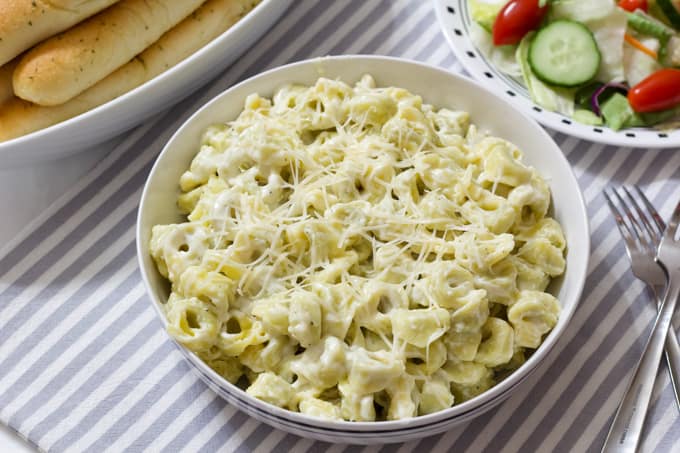 A large bowl of tortellini and part of a salad and bowl of breadsticks. 