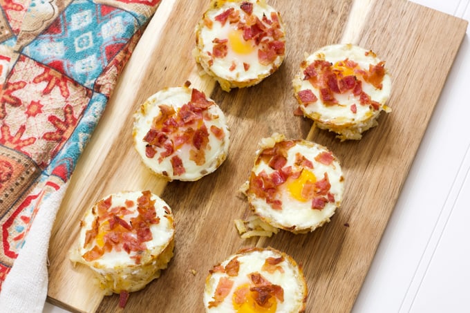 Six eggs in hash brown nests on a wooden cutting board with a multicolored towel next the them.