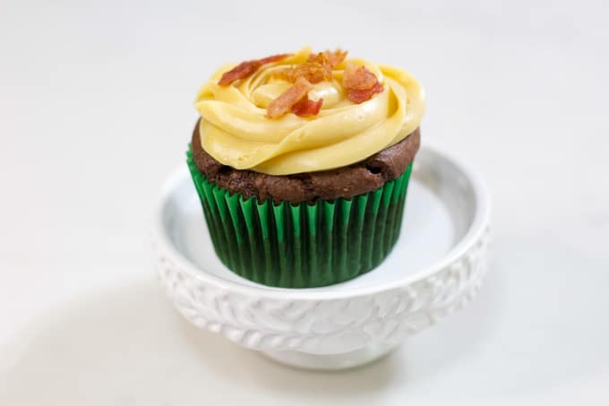 One chocolate cupcake on a white pedestal topped with caramel frosting and bits of bacon.