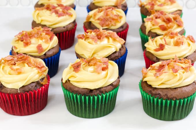 One dozen chocolate cupcakes in colored paper liners topped with frosting and bacon bits.