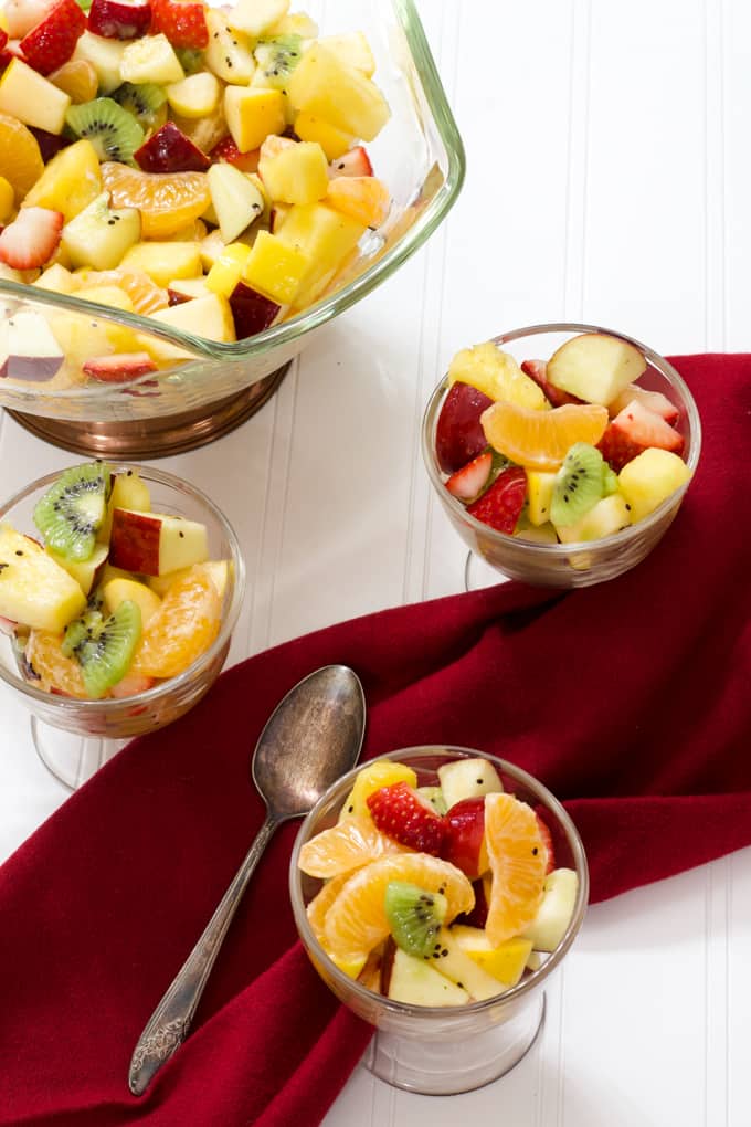 Three small and one large glass bowls of fruit salad with a maroon cloth napkin and spoon on the table.