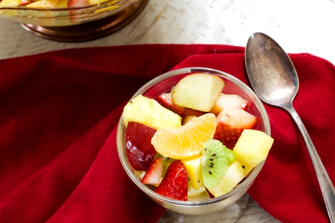Overhead view of one small dessert bowl of fruit salad and a silver spoon on a maroon napkin.