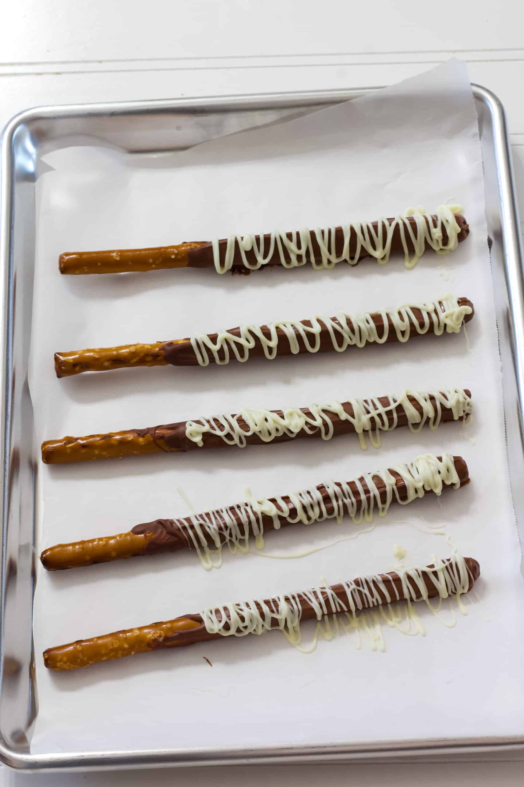 Five pretzel rods that have been dipped in milk chocolate and have a white chocolate drizzle over them. They are laying flat on a silver sheet pan lined with white parchment paper.