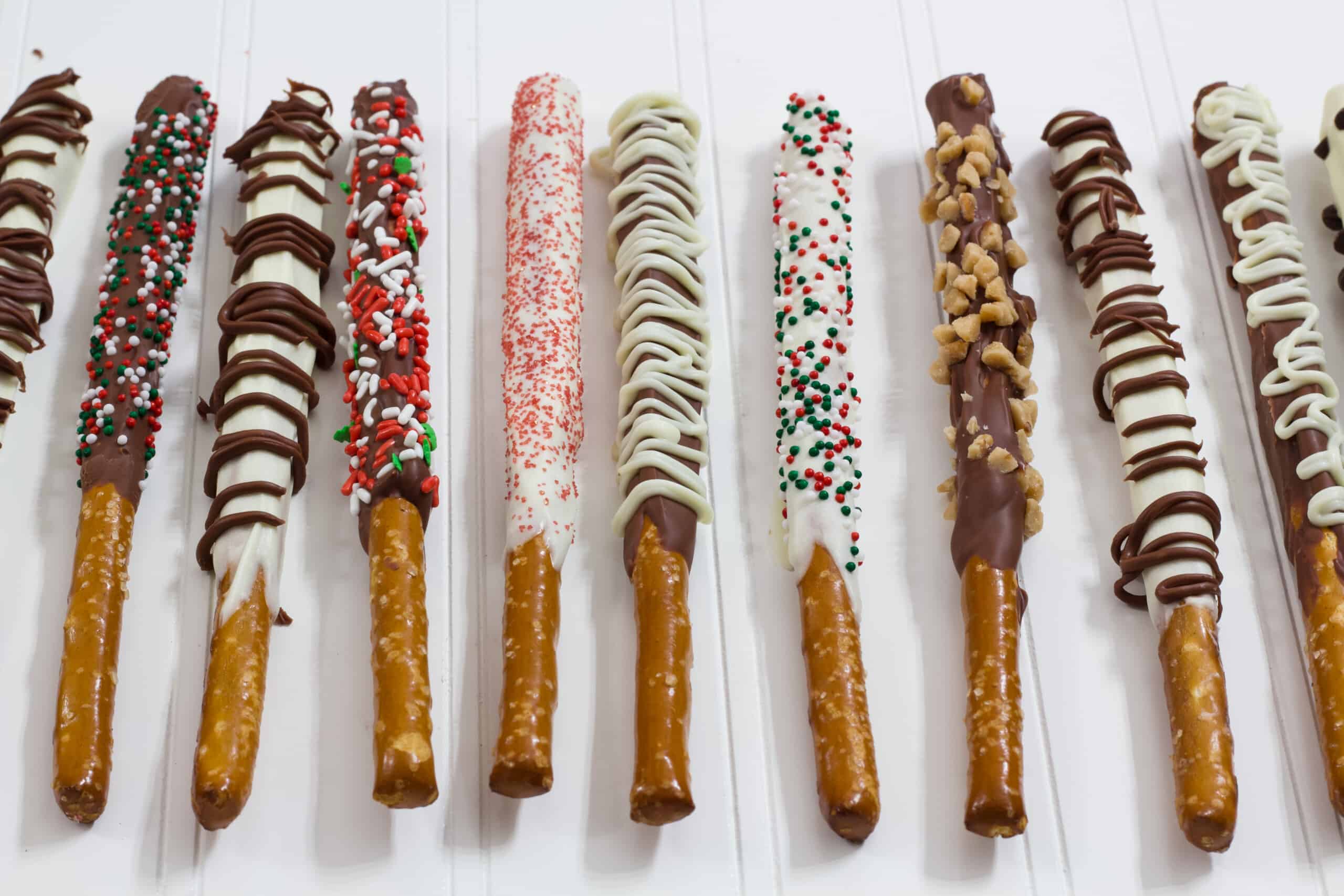 Many chocolate covered pretzel rods laying on a white background. They have various red and green sprinkles on them.