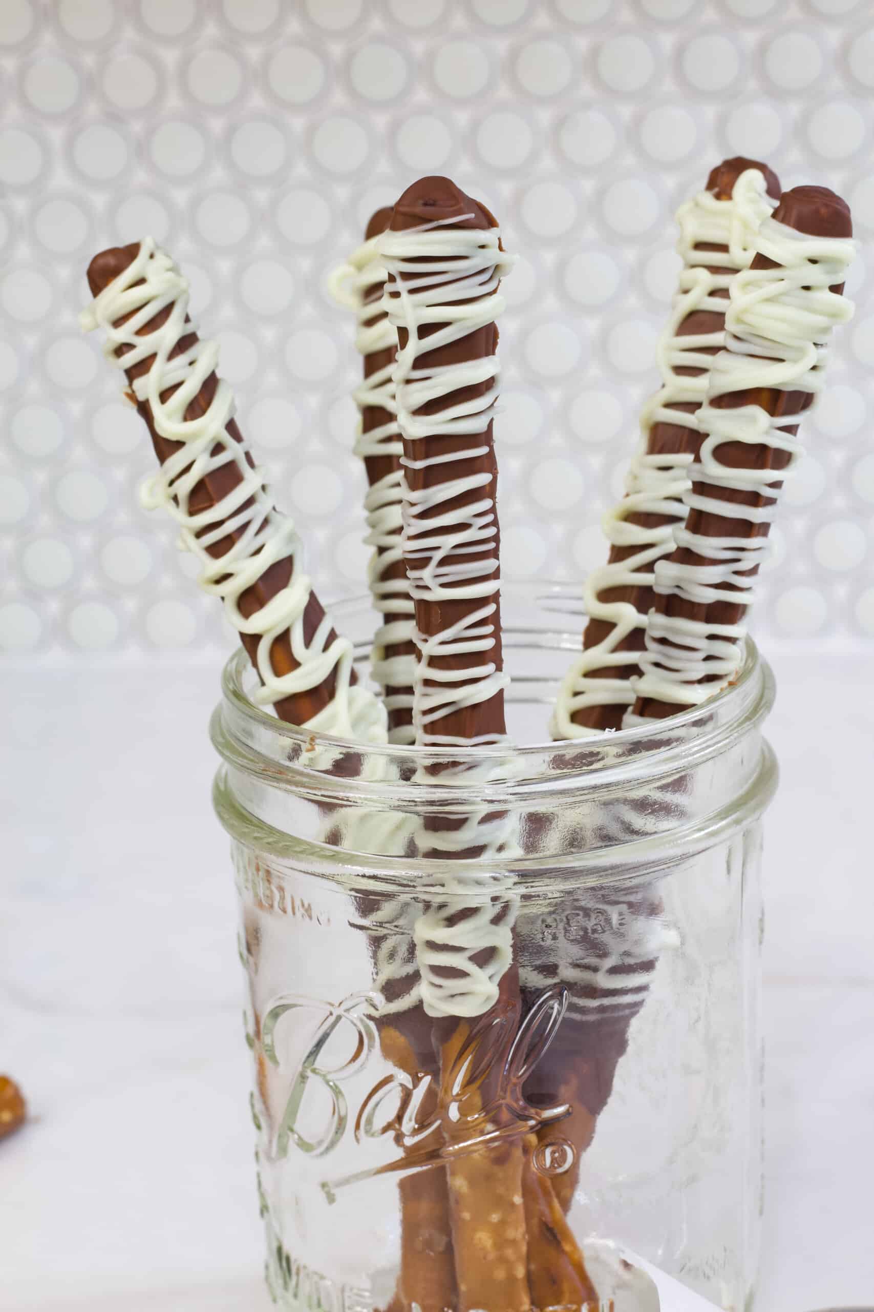 Five pretzel rods that have been dipped in milk chocolate and drizzled with white chocolate in a mason jar.