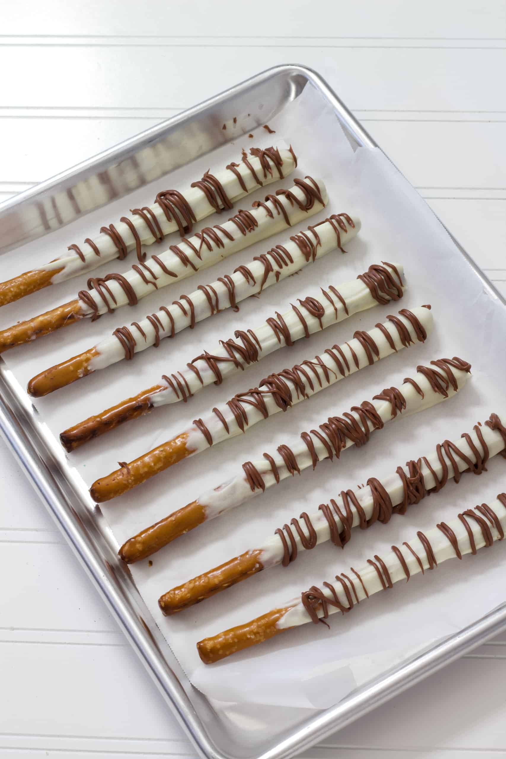 A silver sheet pan lined with white parchment paper that has several pretzel rods that have been dipped in white chocolate and drizzled with milk chocolate.