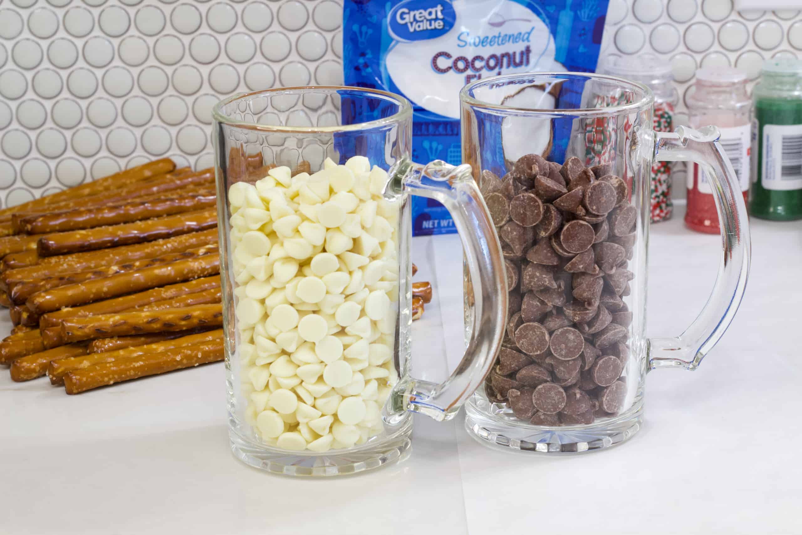 A mug with white chocolate chips and one with milk chocolate chips with pretzel rods in the background.