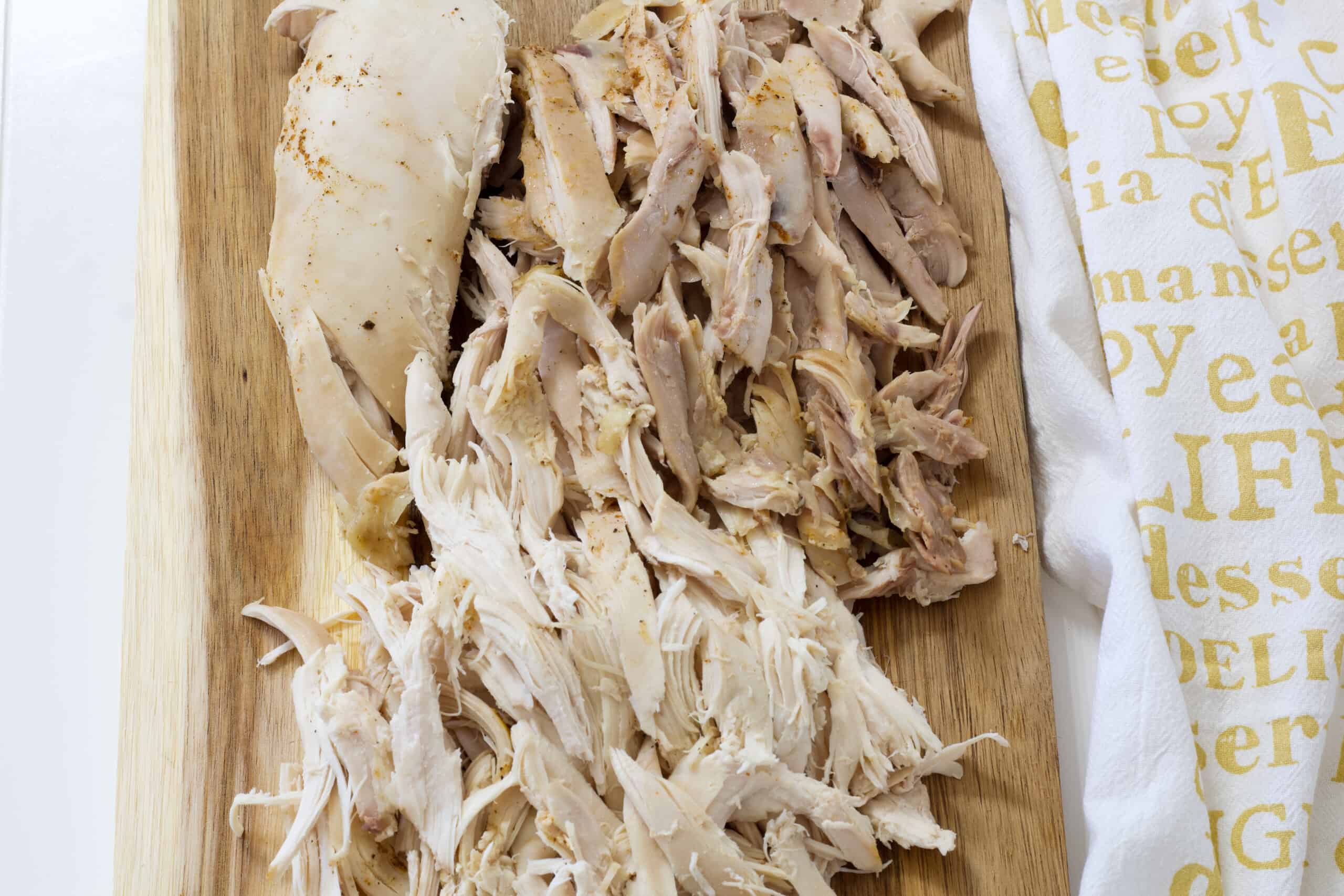 Shredded chicken laying on a wooden cutting board with a tea towel laying next to it.