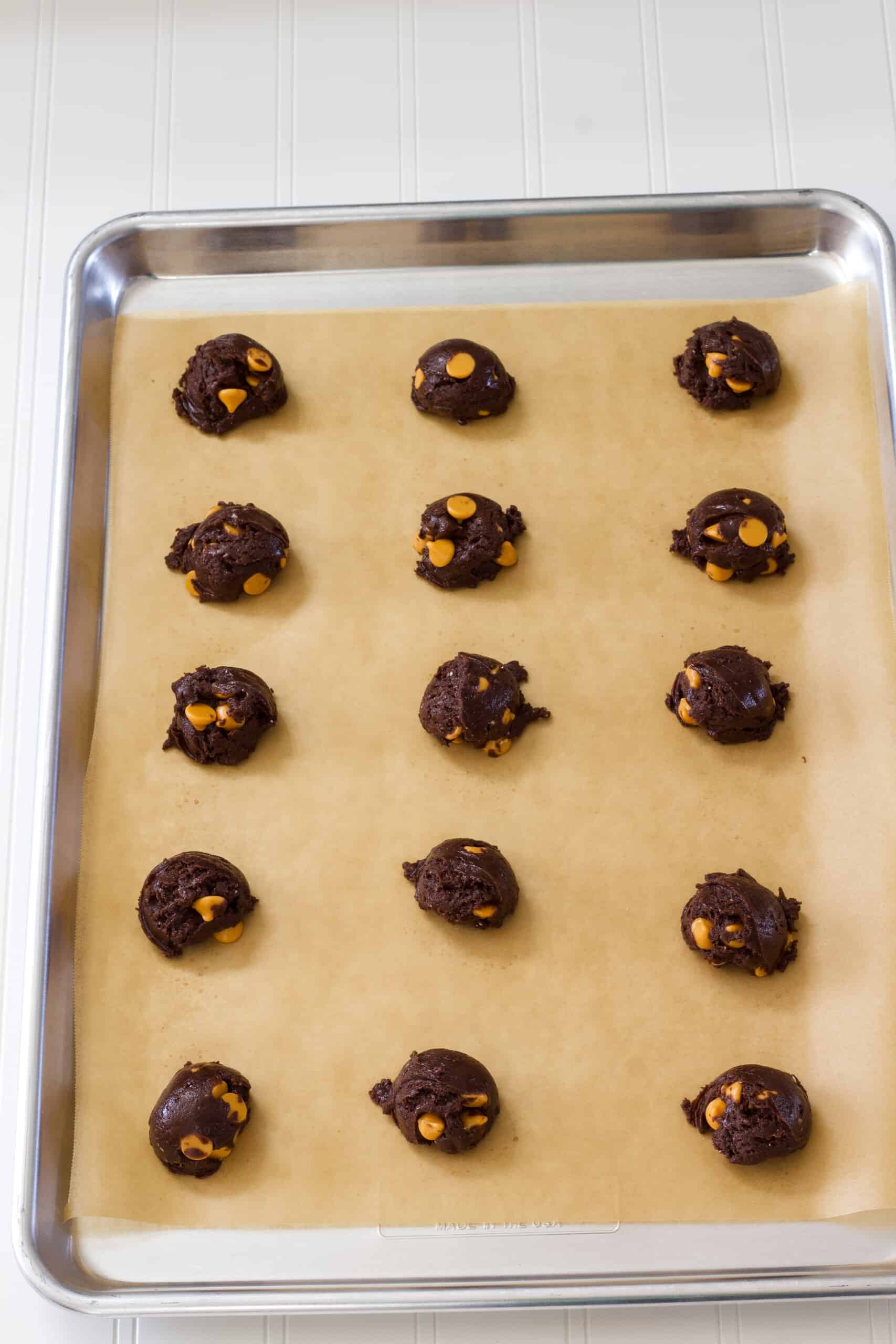 One dozen balls of raw cookie dough on a parchment paper lined baking pan.