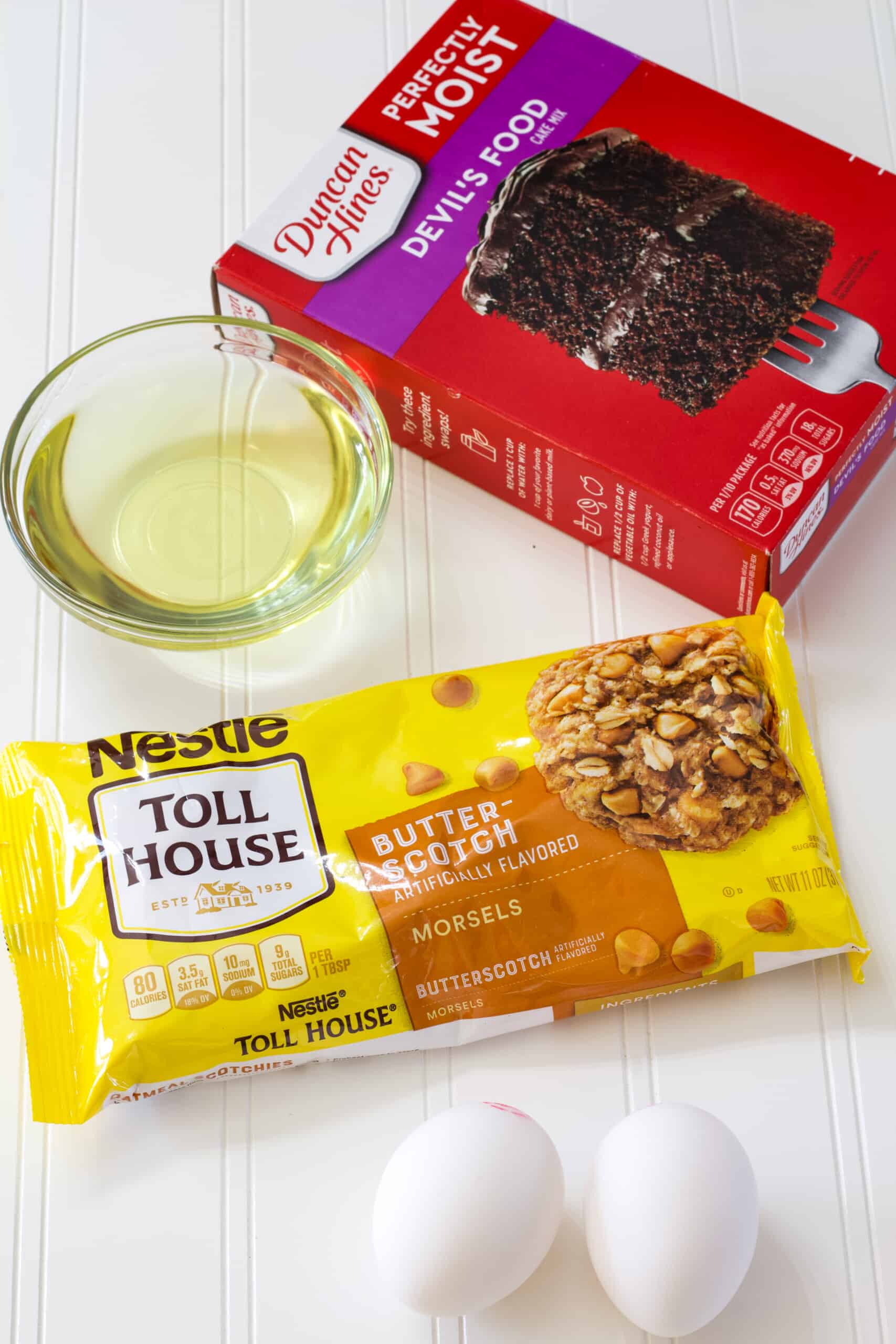 A boxed cake mix, 2 eggs, small bowl of oil and bag of butterscotch chips on the table.