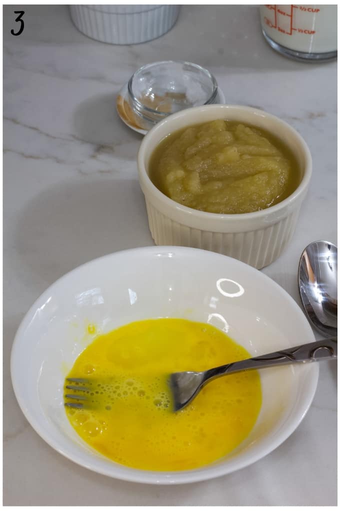 The slightly beaten egg in a small white bowl with a fork in it. The bowl of applesauce is in the background.