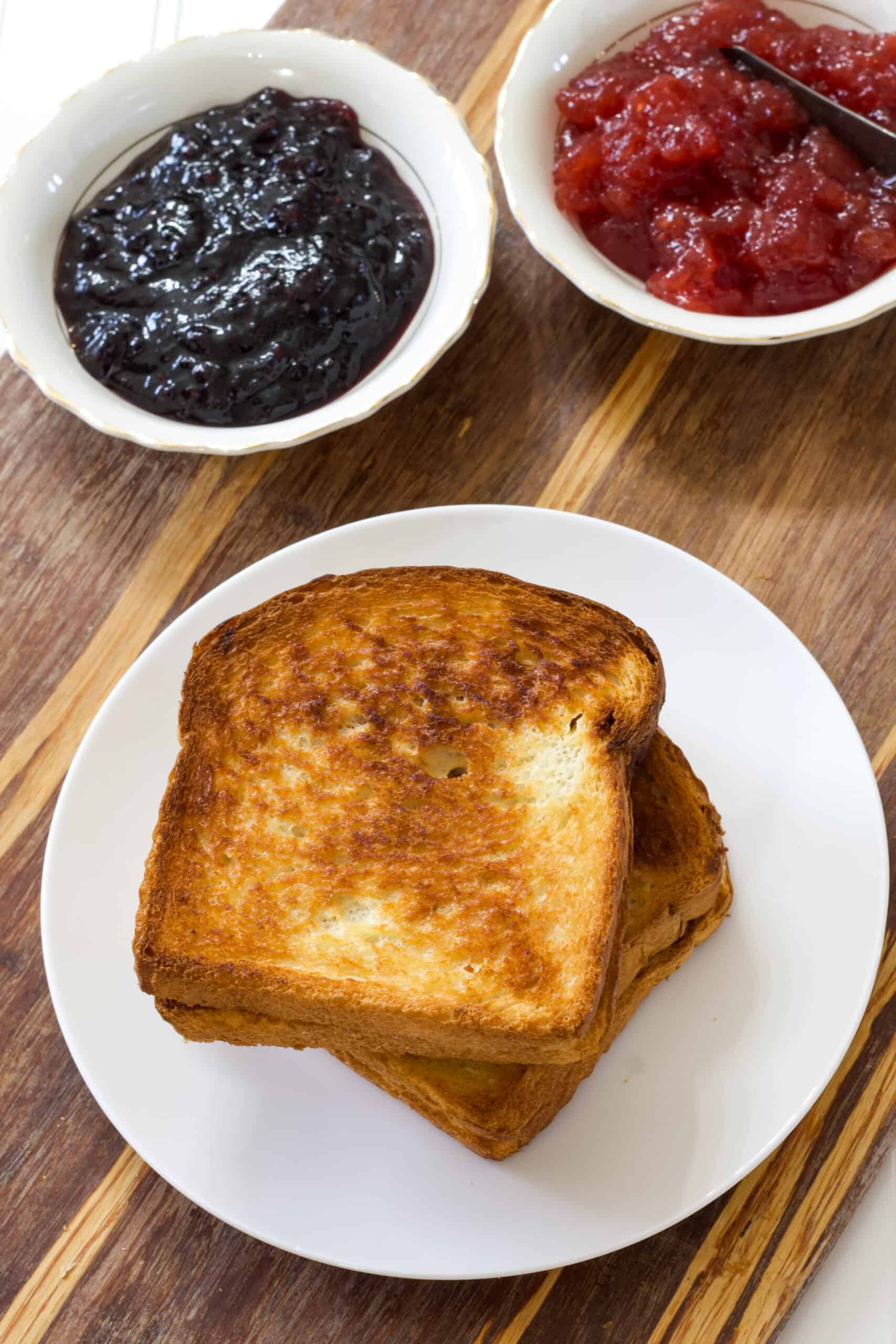Two whole air fried pb & j sandwiches on a white plate with two small bowls of jelly in the background.