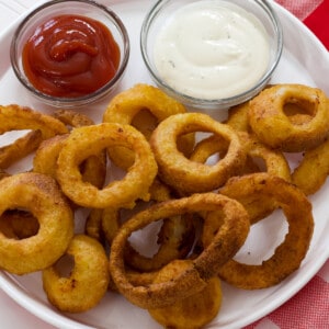 The feature image of the onion rings on a white plastic plate with a small bowl of ranch dressing and a small bowl of ketchup on it too.