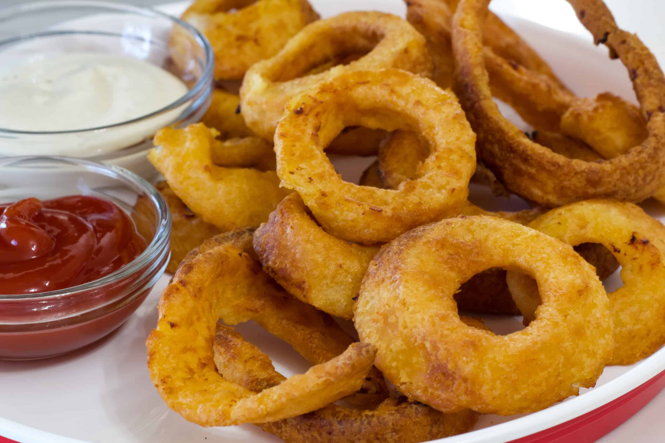 Very close shot of the air fried onion rings on plate with a small bowl of ketchup and ranch dressing sitting next to them.