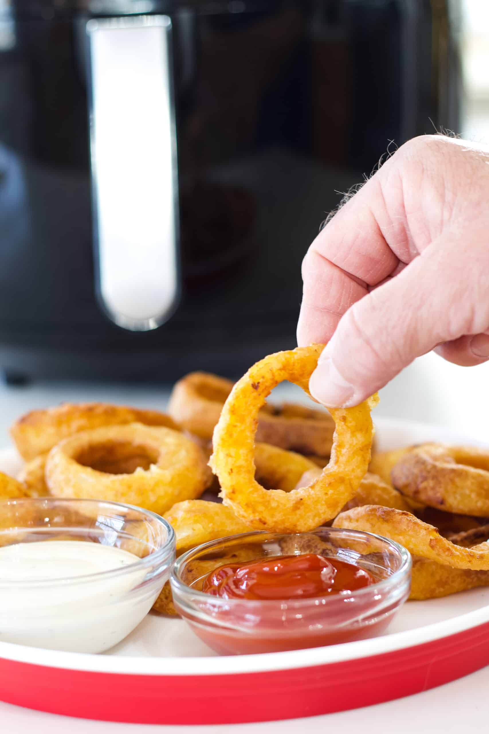 A hand holding an onion ring to dip into a bowl of ketchup.