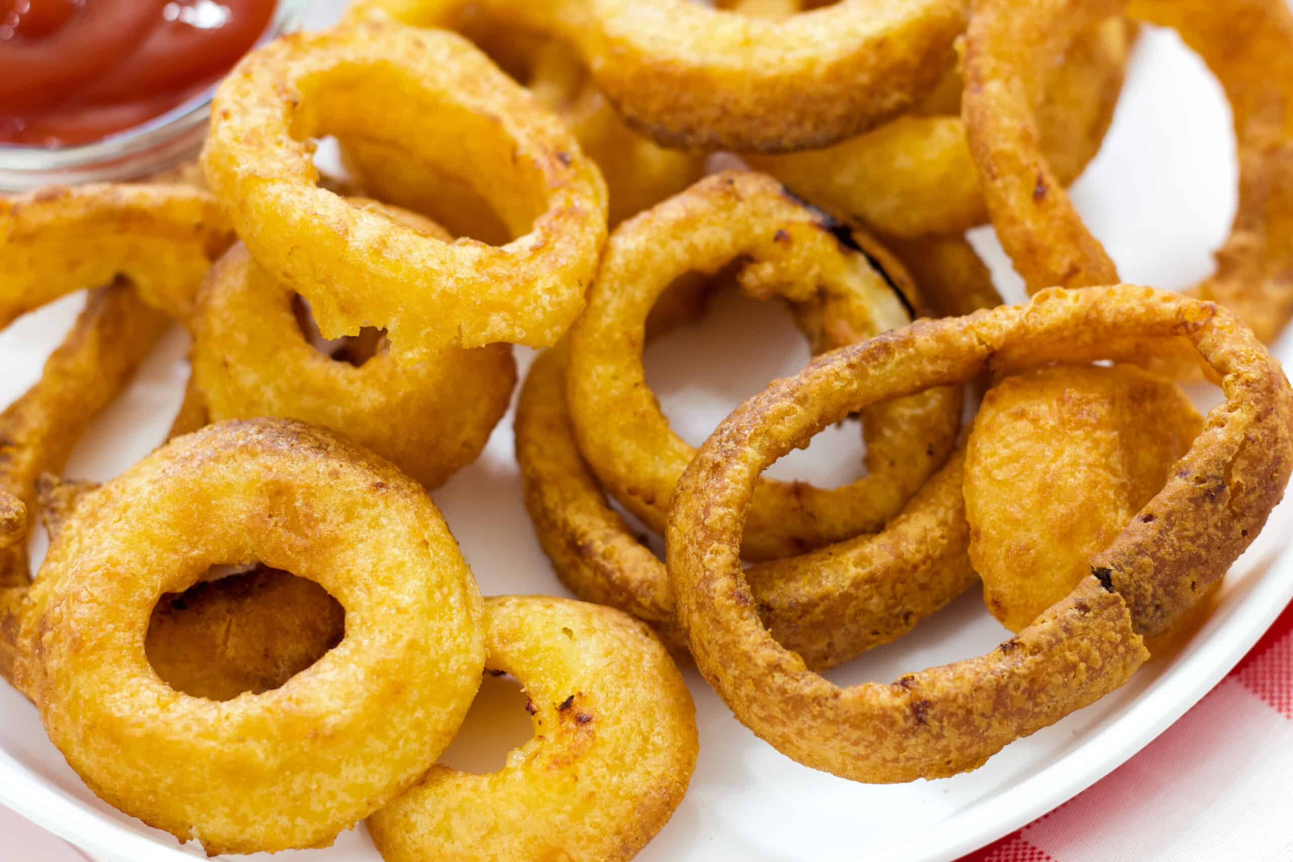 close up of the onion rings on a white plastic plate with a bowl of ketchup in the top left corner of the image.