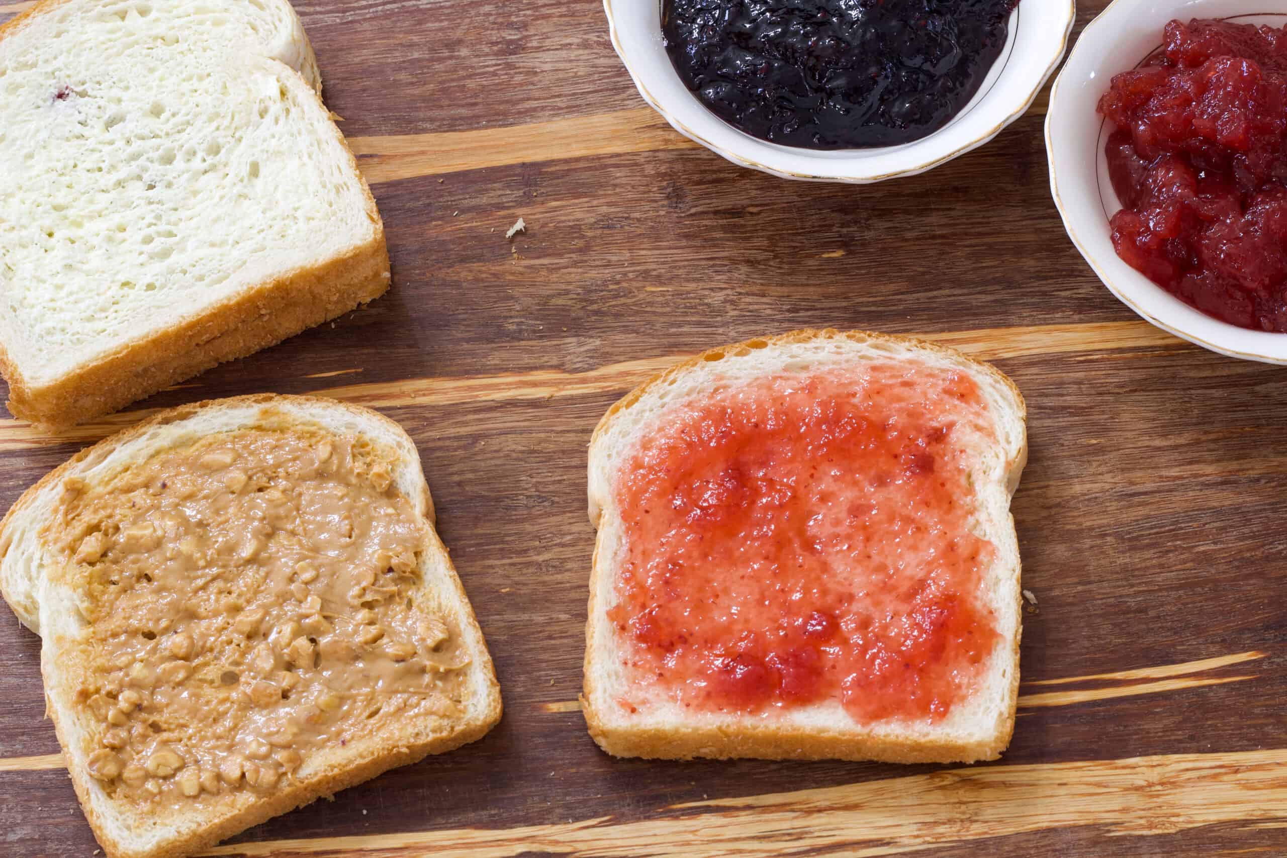 Bread on a wooden cutting board, one with peanut butter and one with jelly and a whole sandwich and two small bowls of jelly.