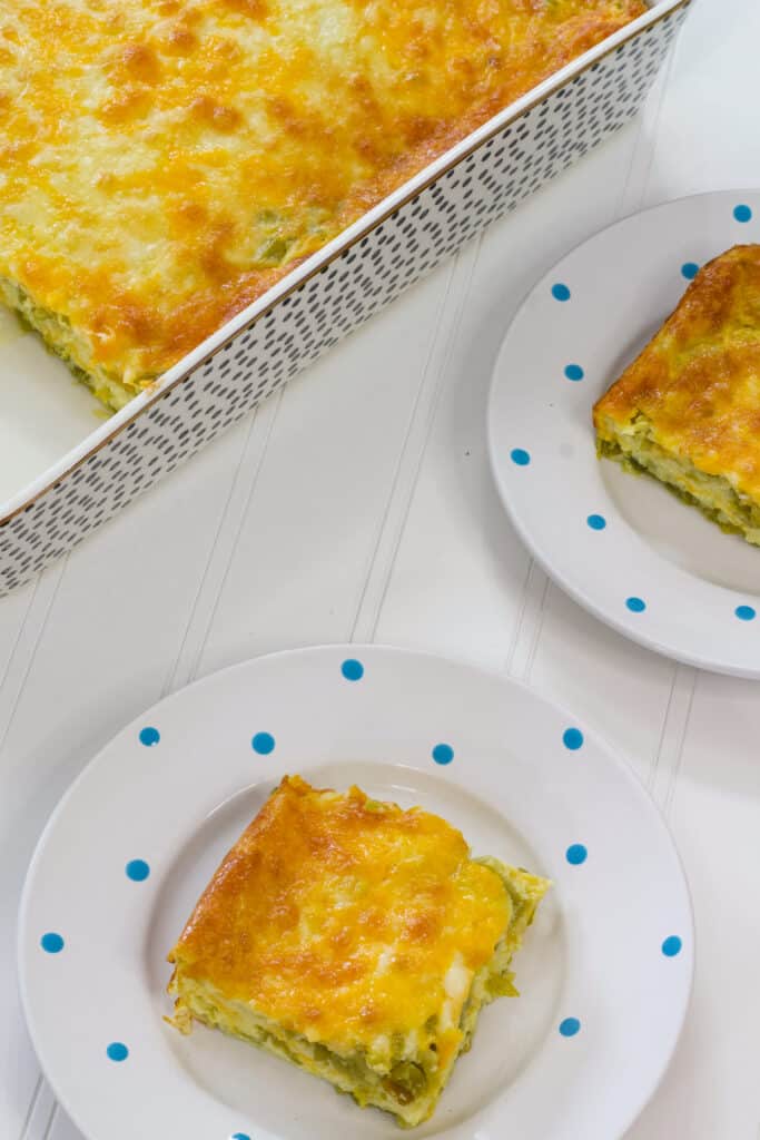 A serving of chile rellenos casserole on a plate with a portion of the remaining casserole and one other serving in the shot.