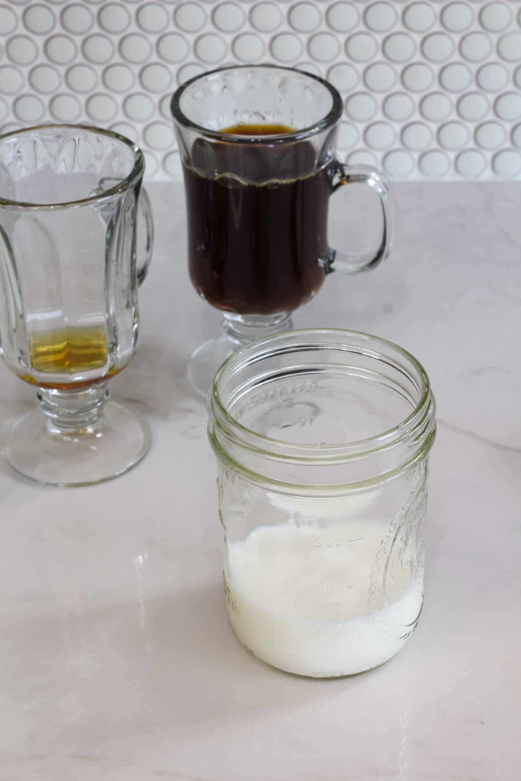 A cup of black coffee and a cup with maple syrup in it in the background and a mason jar with a little milk in it in the foreground.