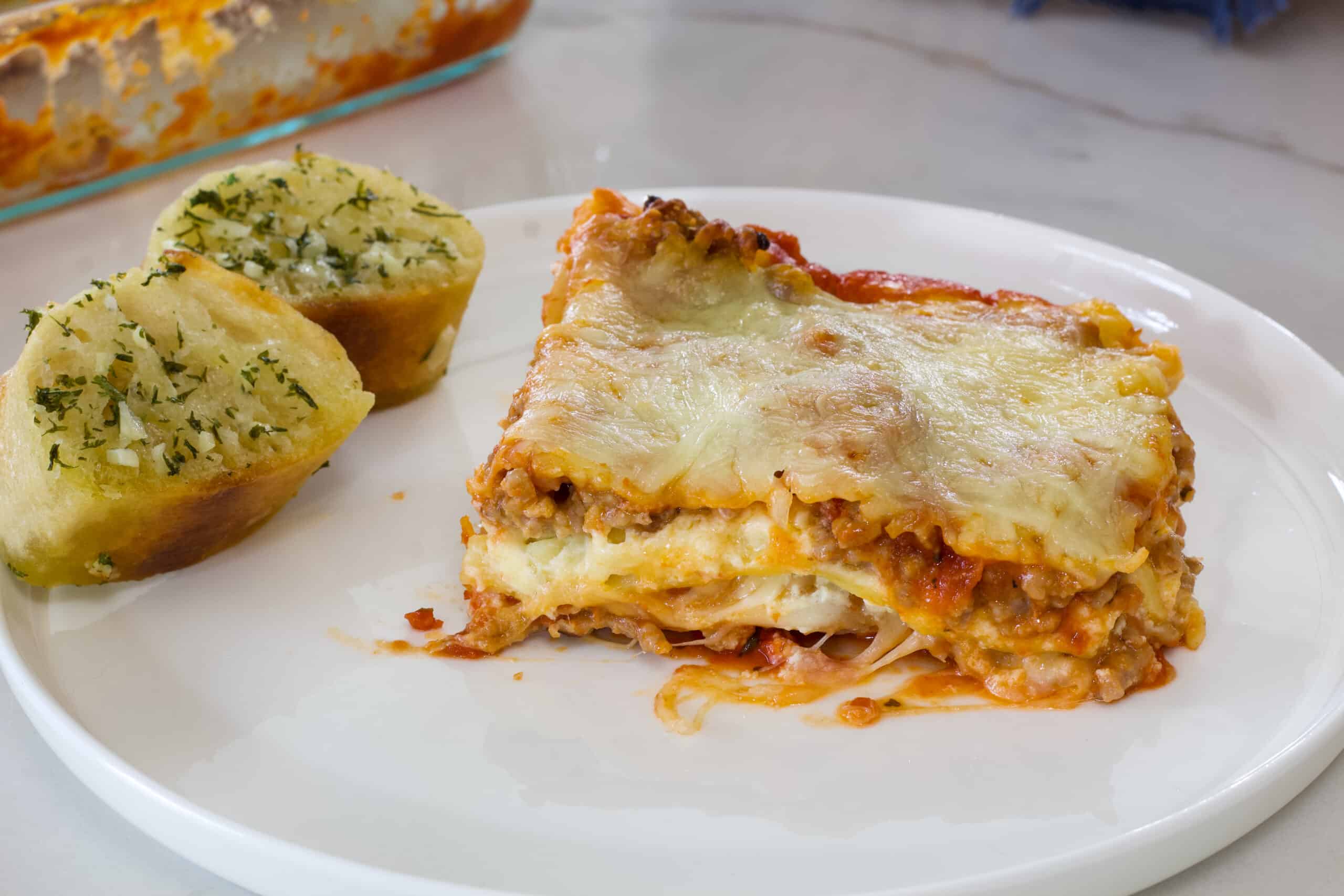 One serving of lasagna and two pieces of garlic bread on a white plate.