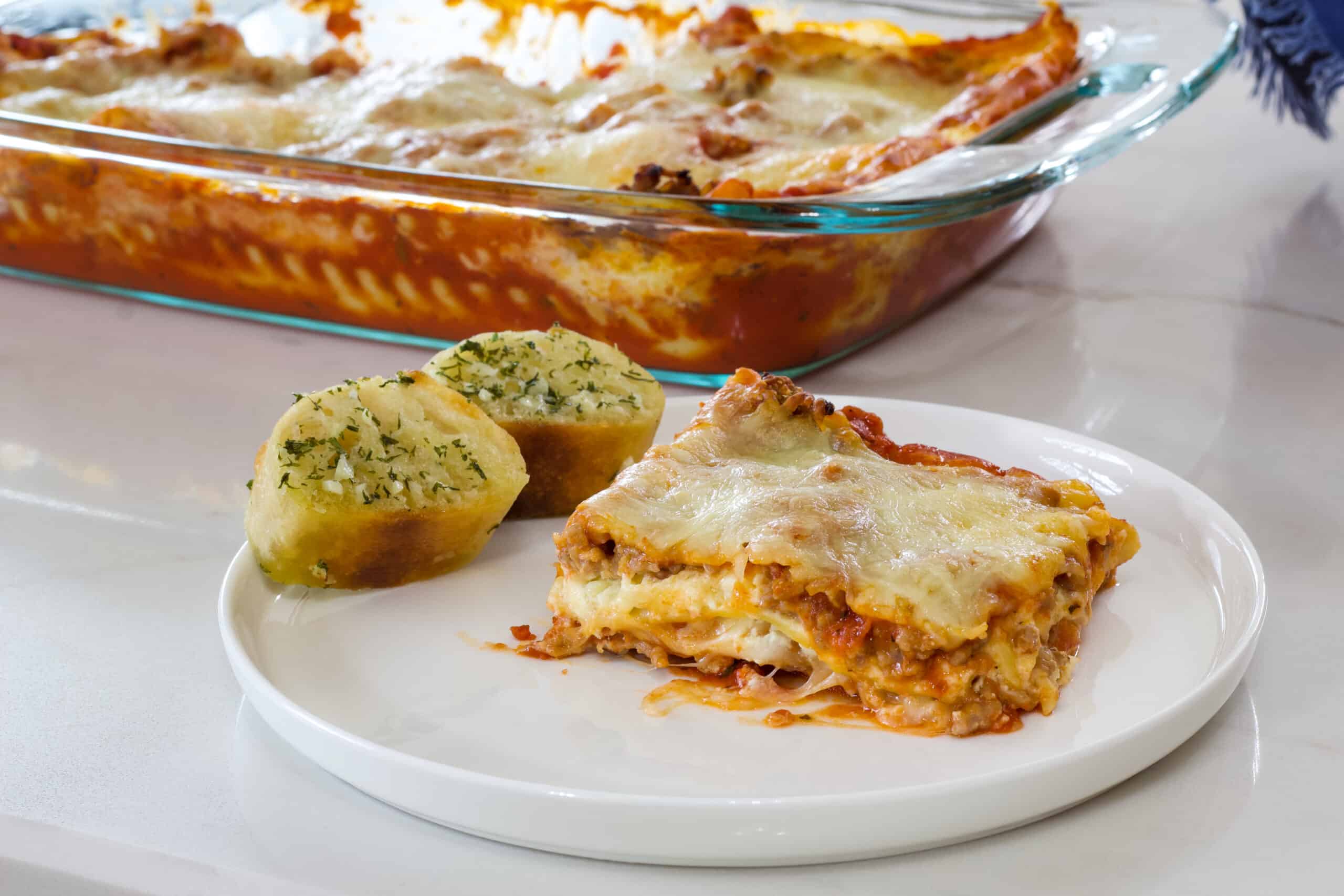 One serving of lasagna and two pieces of bread on a plate and the pan of lasagna in the background.