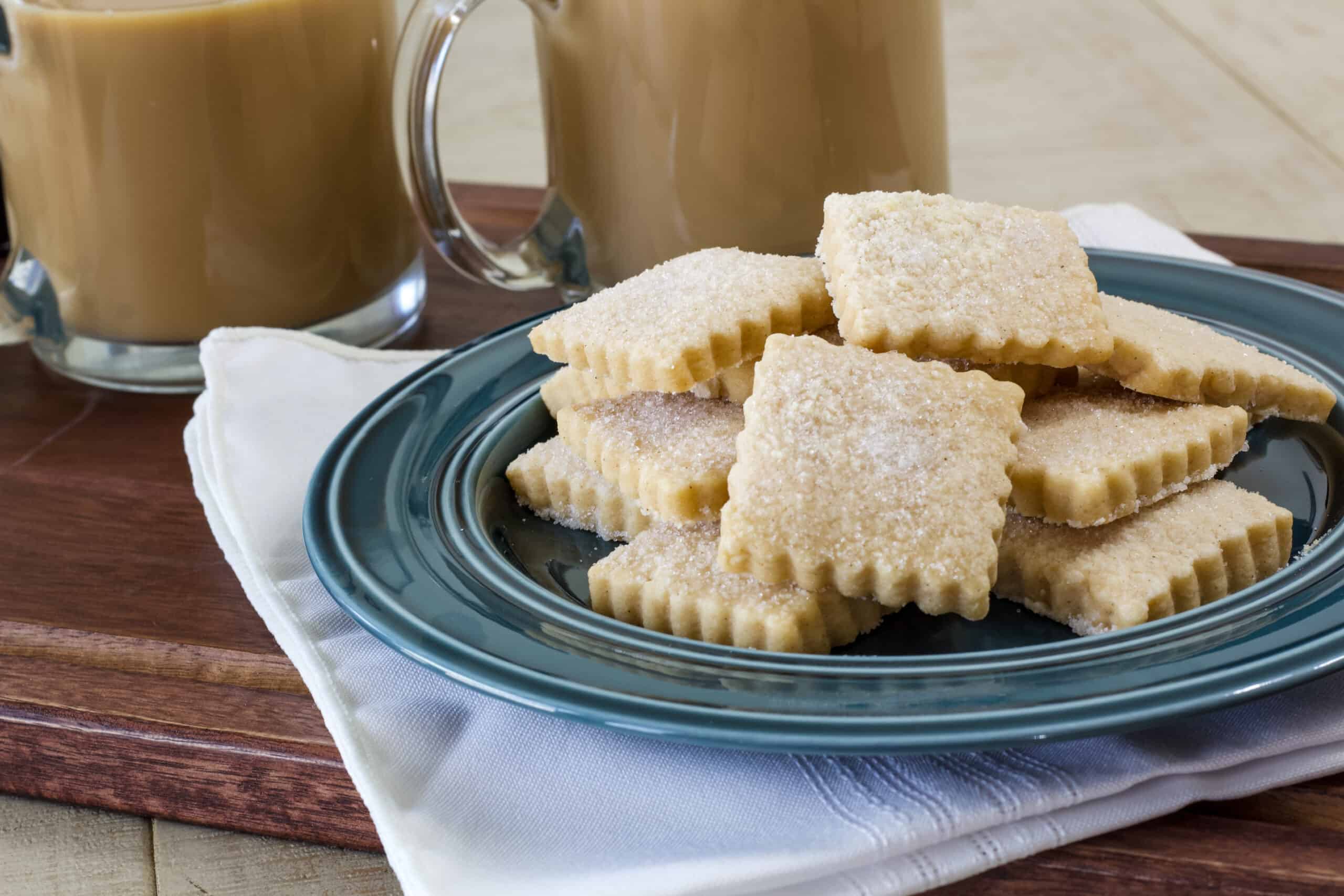 A plate of square cookies on a plate that is on a wooden serving tray with two cups of coffee.