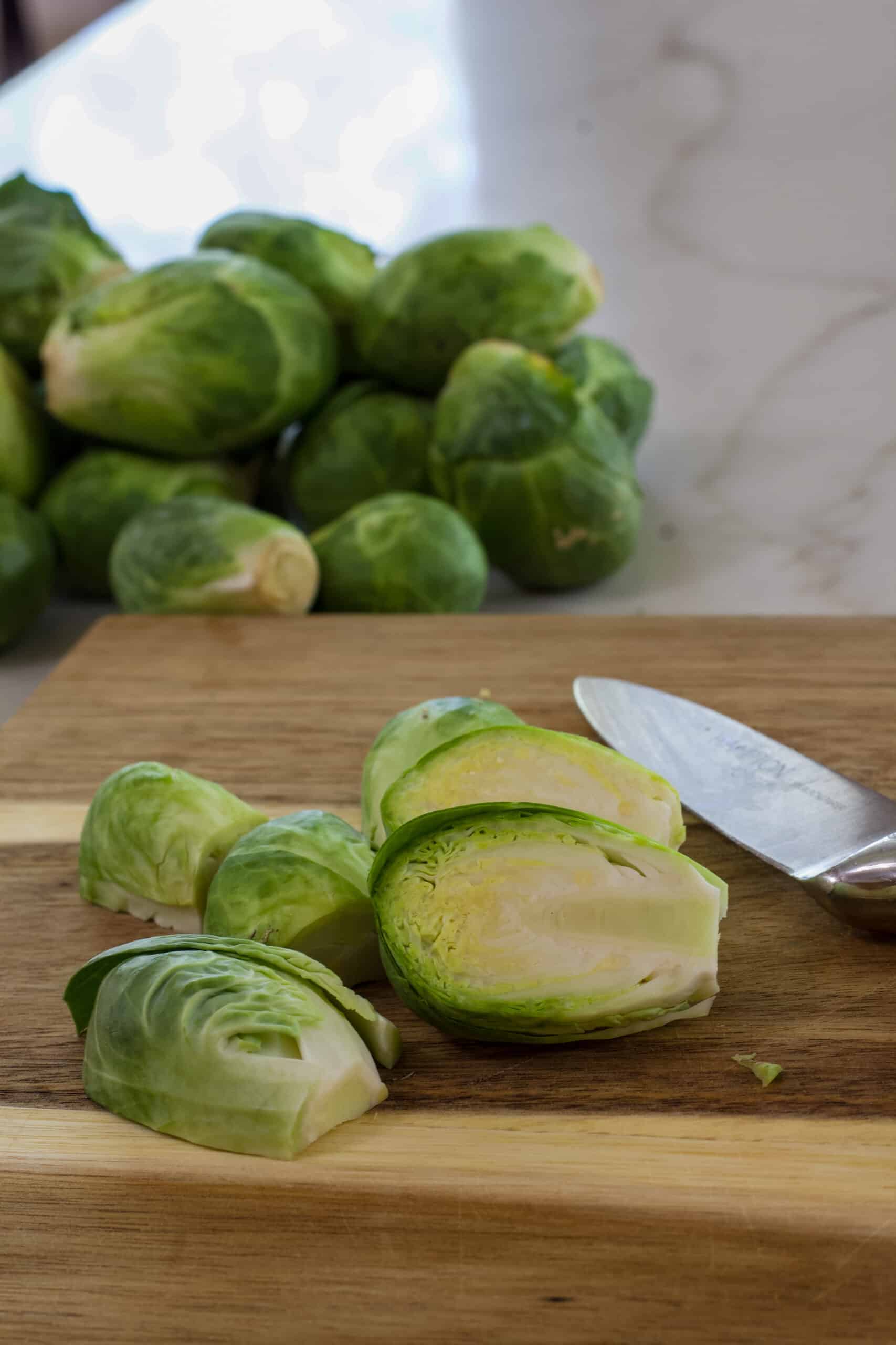 Several but Brussels sprouts on a wooded cutting board with a pile of sprouts in the background.