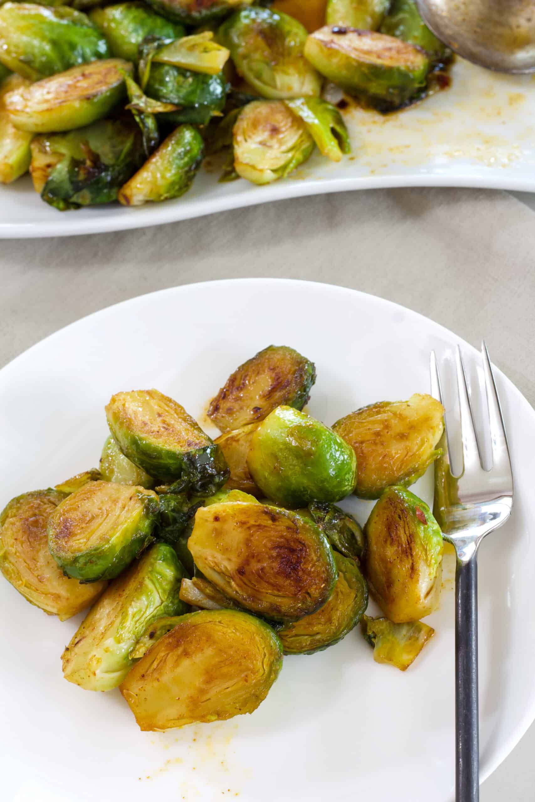 A white plate with a serving of Brussels sprouts on it in the foreground and a plate of brussels sprouts in the background.