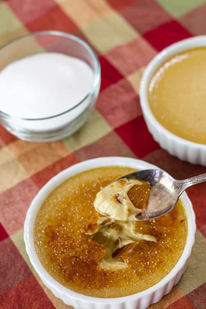 A ramekin of creme brulee without sugar in the background and one with the sugar topping in the foreground that has a spoonful above it.
