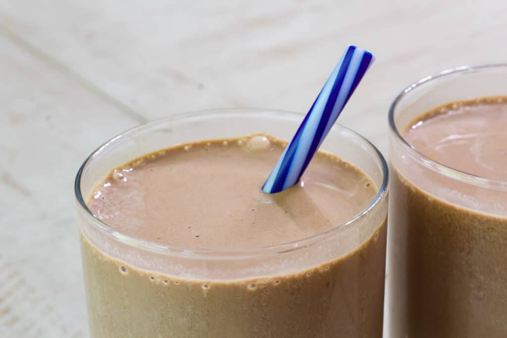 Close up view of the top of one smoothie in a glass with a blue striped straw sticking out of it.