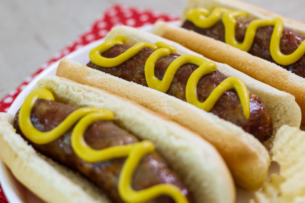 Three cheddar brats in hot dog buns with mustard on them.