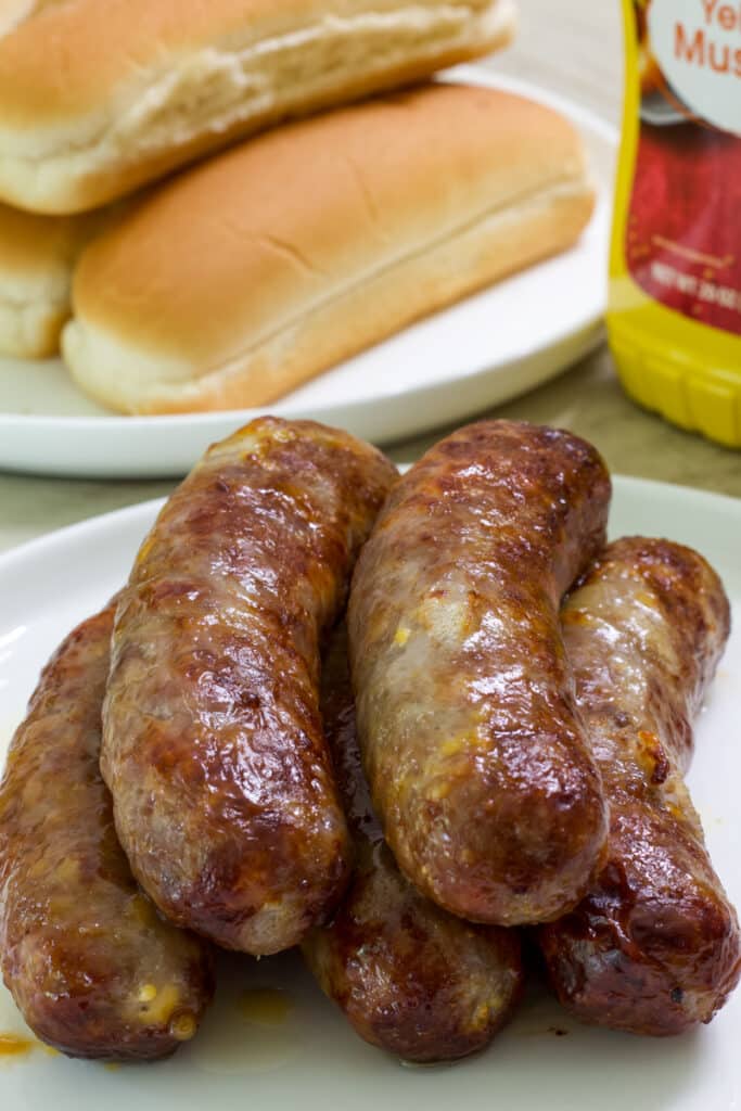 Five cooked cheddar brats on a plate in the foreground and buns and a bottle of mustard in the background.