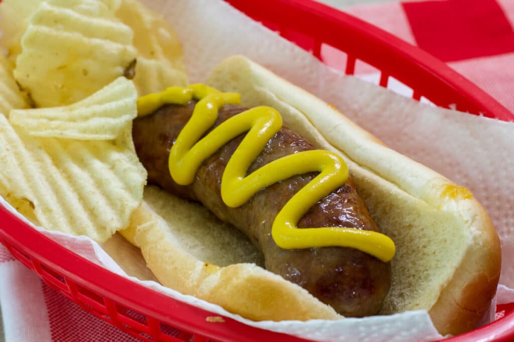 One cheddar bratwurst in a hot dog bun with some chips in a red basket.