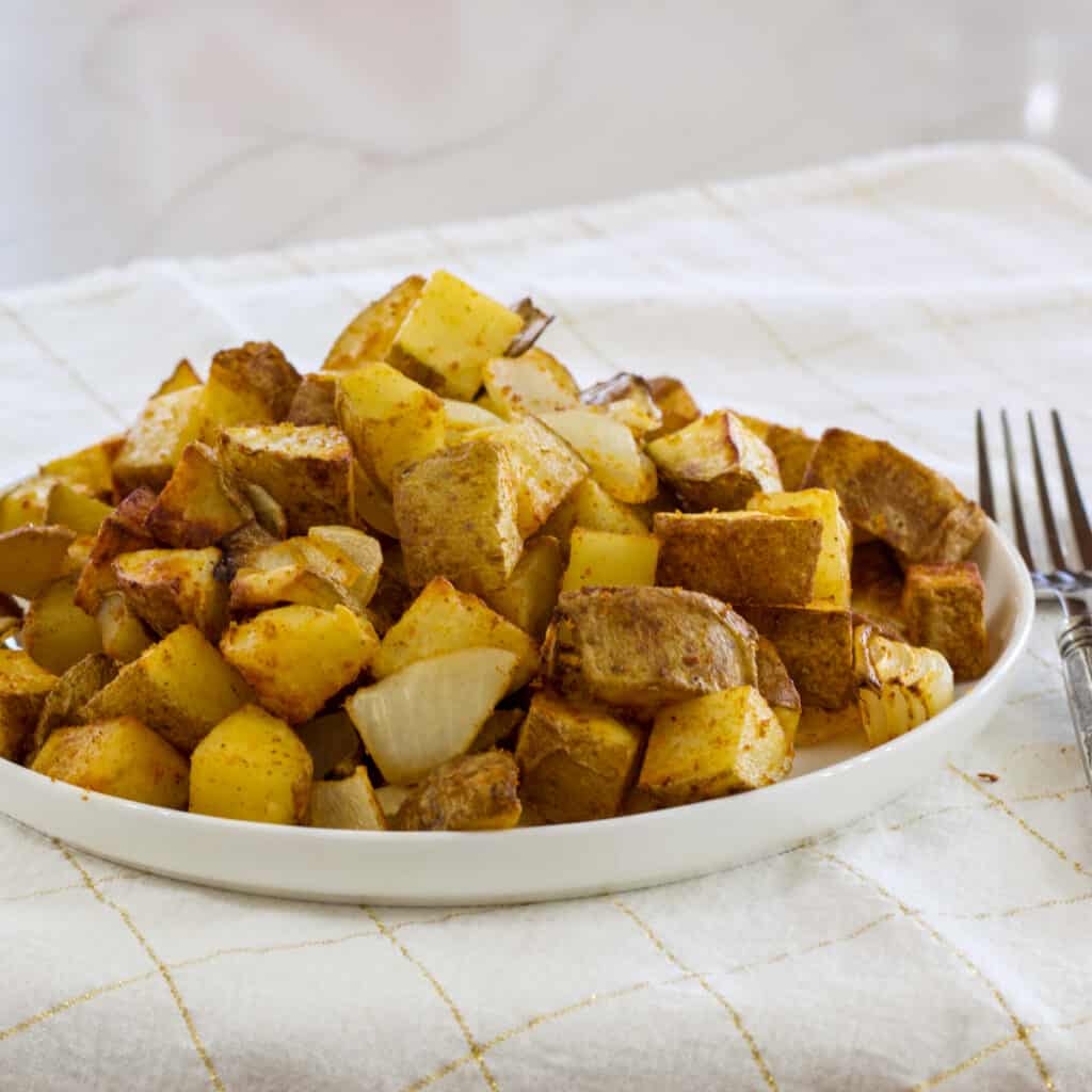 The plate of potatoes and onions on a white and gold napkin with a silver fork on the right side.