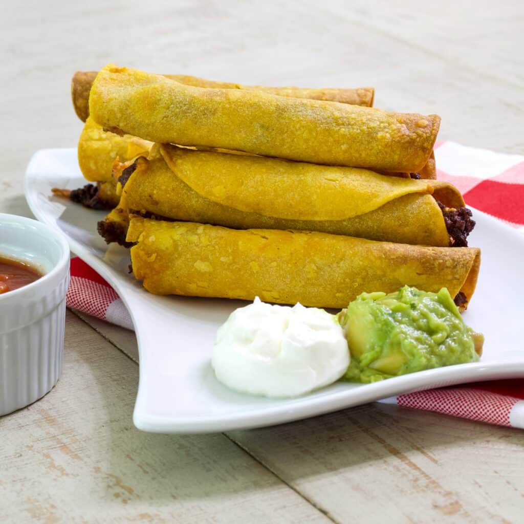 Several beef taquitos and a scoop of sour cream and guacamole on a plate and you can see part of a bowl of salsa on the left.