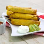 Several beef taquitos and a scoop of sour cream and guacamole on a plate and you can see part of a bowl of salsa on the left.