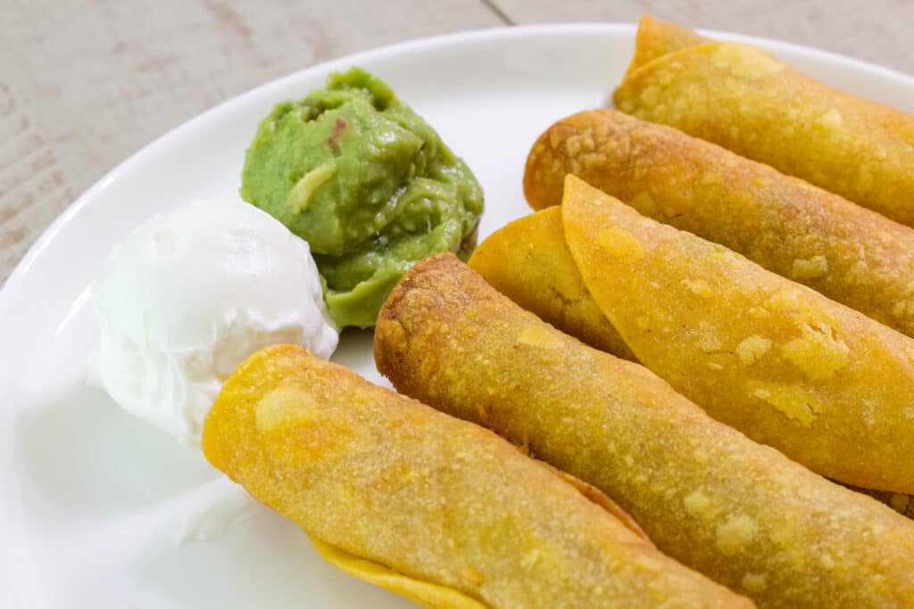 A close up of just the end of five beef taquitos with a scoop of sour cream and guacamole on a white plate.