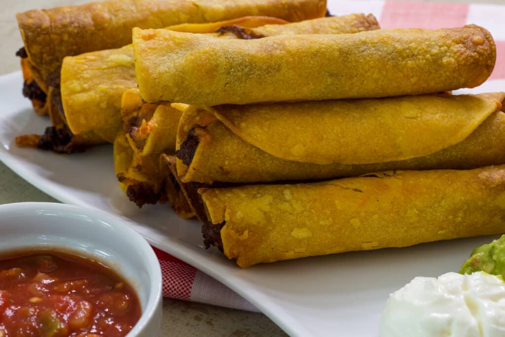 Several beef taquitos on a white plate with a small bowl of salsa in the corner of the photo.