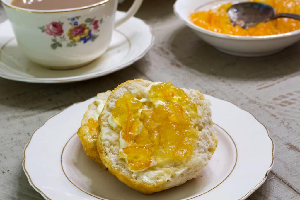A cut biscuit on a plate with butter and orange marmalade on it.