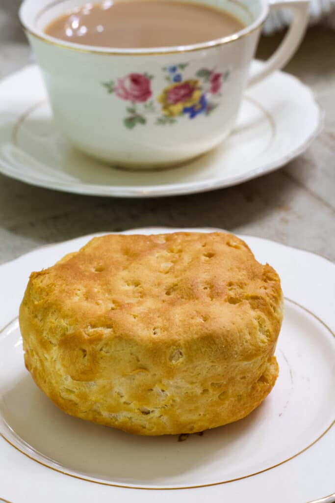 One whole biscuit on a plate in the foreground and a cup of coffee in the background.