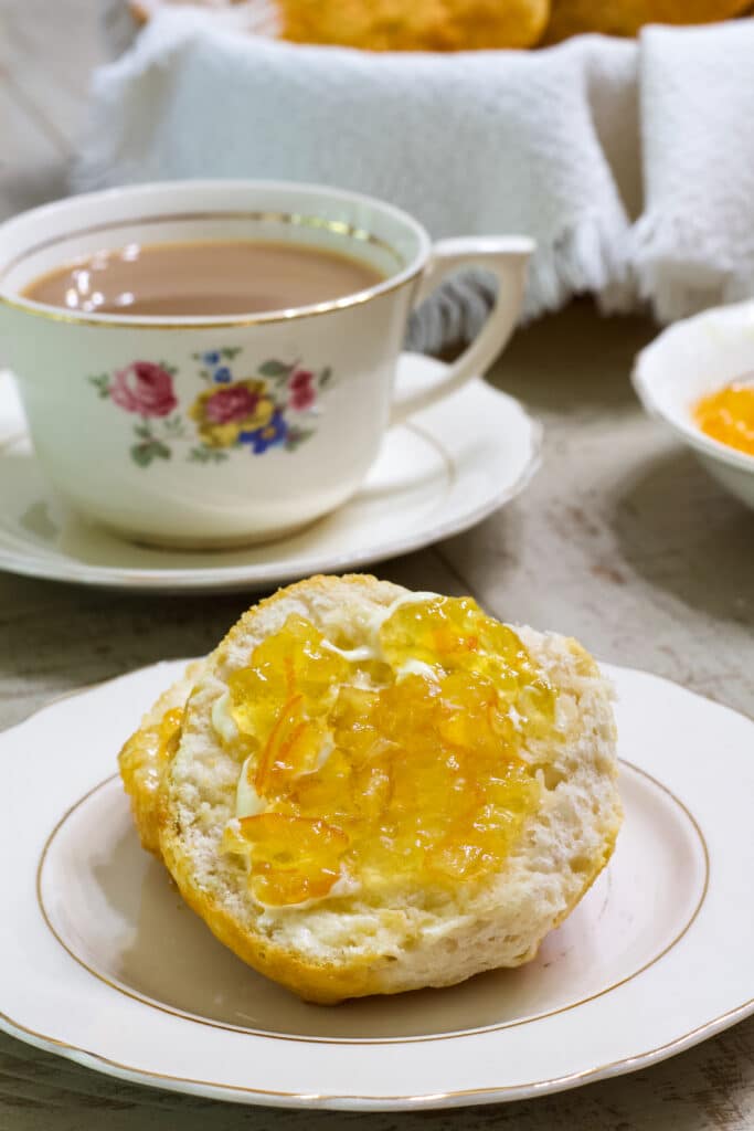 A cut biscuit with butter and orange marmalade on it in the foreground with a cup of coffee and small bowl of marmalade in the background.
