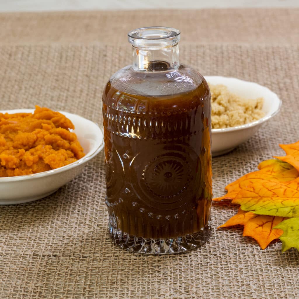 A jar of syrup and a small bowl with pumpkin puree and a small bowl of brown sugar on a burlap background.