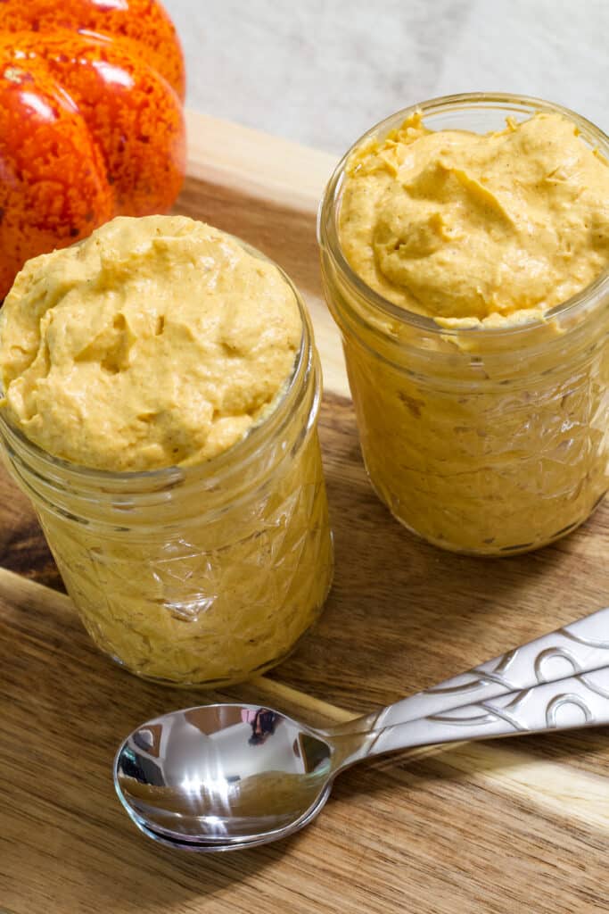 Two small mason jars filled with pumpkin fluff, there are two spoons next to them and a pumpkin in the background.