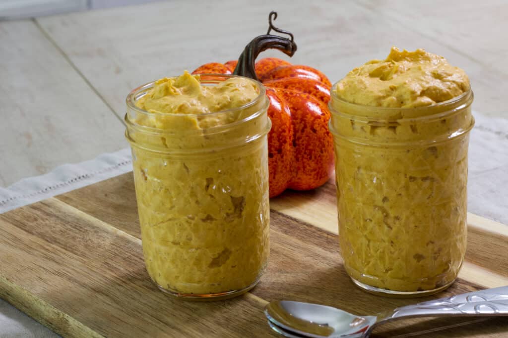 Two small mason jars filled with pumpkin fluff, two spoons in front of them and an artificial pumpkin behind them.