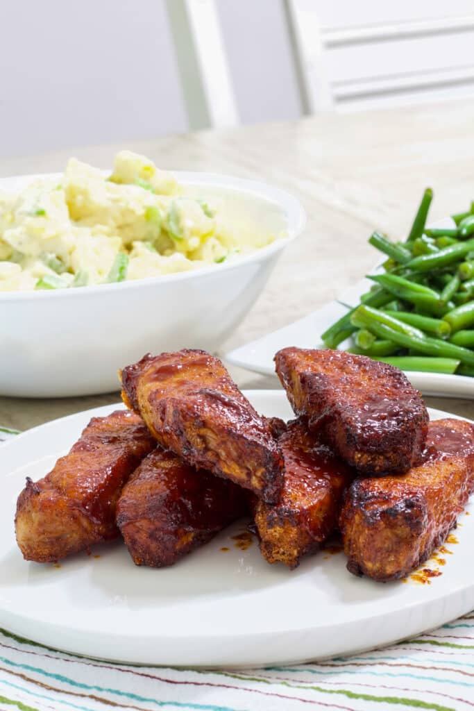 A plate of ribs in the foreground and a bowl of potato salad and plate of green beans in the background.