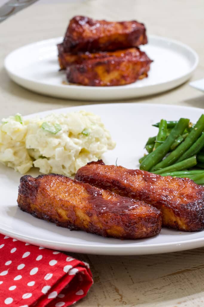 Two ribs on a plate with potato salad and green beans, there isa red an white napkin sitting next to the plate.