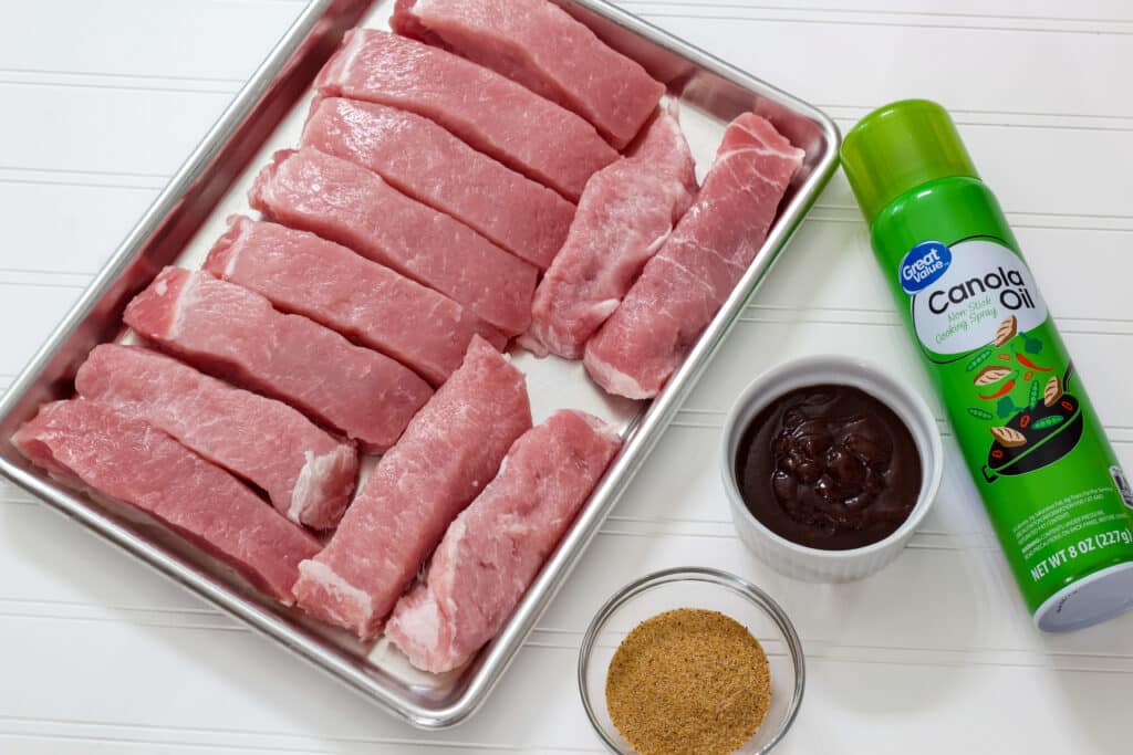 Uncooked ribs on a sheet pan, dry rub and barbecue sauce in small bowls and a can of cooking spray on a white background.